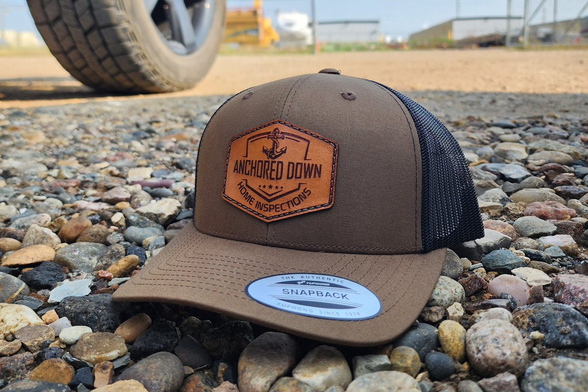 Brown trucker cap with an engraved Anchored Down leather patch on gravel