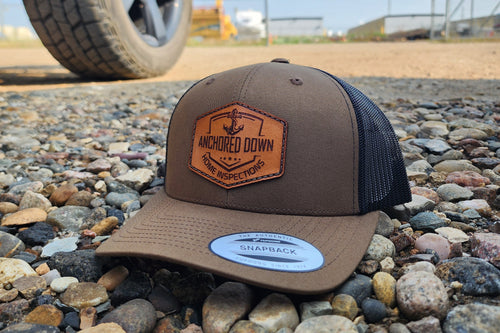 Brown trucker cap with an engraved Anchored Down leather patch on gravel