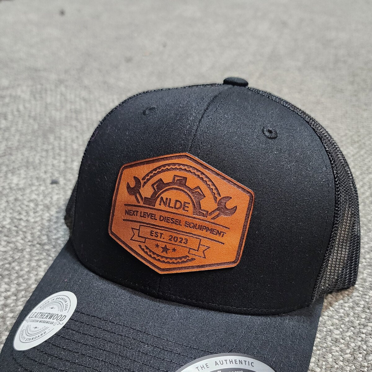 Hexagon leather patch cut to shape with a gear and wrench logo, engraved Next Level Diesel Equipment Est. 2023, sewn on a black trucker cap