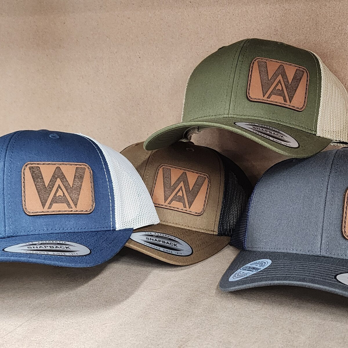 Four trucker caps in navy, brown, olive and grey, each with the same engraved WA leather patch sewn on