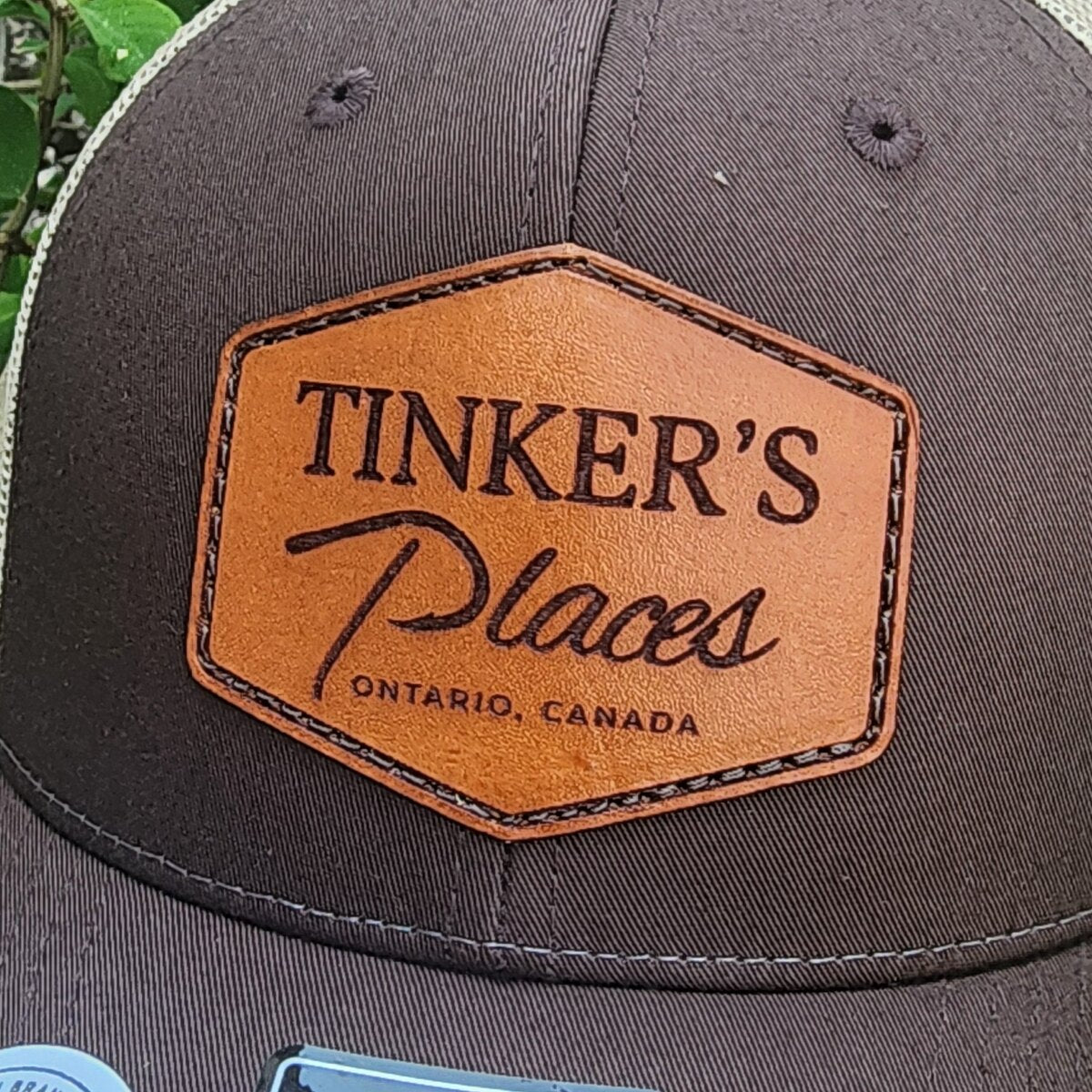 Close-up of a tan hexagon leather patch engraved Tinker's Places Ontario, Canada with a stitched border sewn onto a brown trucker cap